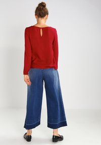 Rode longsleeve top met een keyhole-detail aan de achterkant, gecombineerd met brede blauwe denim broeken met een rafelige onderrand en subtiele stiksels. Zwarte flats.