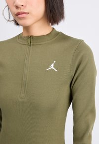 Alyvuogių spalvos cannelé ilga rankovė palaidinė su aukštu apykakle ir ketvirčio užtrauktuku. Kairiajame krūtinės pusėje yra balta Jumpman logotipas.