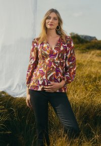 Kleurige, gebloemde blouse met een diepe V-hals en puffmouwen, gedragen met een donkere spijkerbroek. Gekozen in een grasveld met een witte achtergrond.