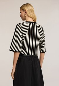 Vrouw met blond haar, draagt gouden ringoorbellen en een zwart-wit gestreepte korte mouw top, gecombineerd met een zwarte rok.