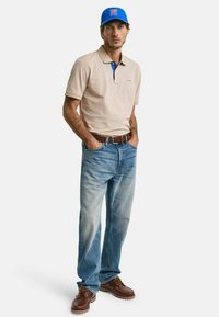 Beige polo skjorte med blå krage, lyseblå jeans, brune snøring sko. Modellen står med hendene i lommene, iført en blå baseballcaps.