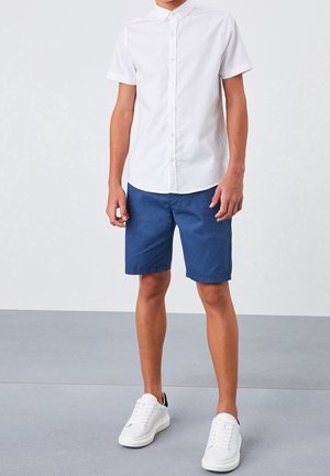 Personne portant une chemise blanche à manches courtes boutonnée, un short bleu jusqu'aux genoux et des baskets blanches, debout sur un sol gris.