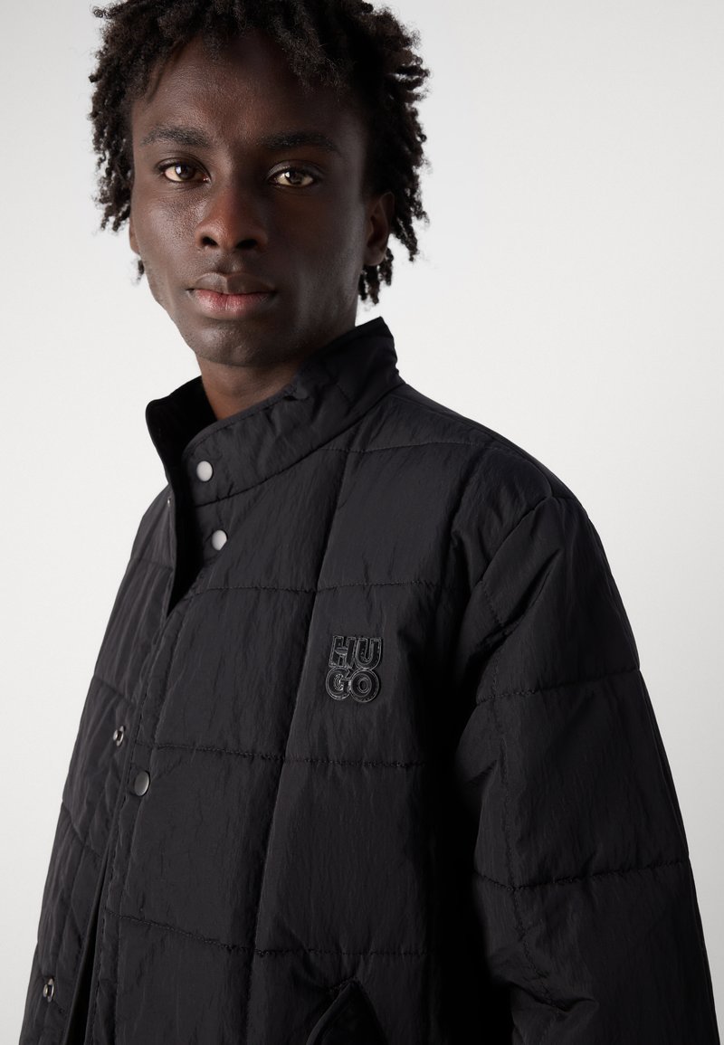 Veste noire matelassée avec une fermeture à boutons-pression, dotée d'un col structuré et d'un logo "HUGO" brodé sur la poitrine.