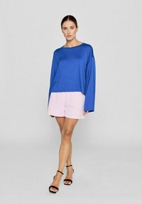 Blå langærmet bluse med en afslappet pasform, parret med lyserøde shorts. Shortsene er enkle med en glat tekstur. Sorte sandaler med stropper.