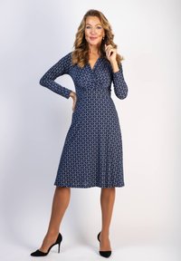 Mekko tummansinisestä kankaasta, jossa on pitkät hihat, muodikas yl�äosa ja leveähelma. Siinä on valkoinen geometrinen kuvio ja V-pääntie. Yhdistetty mustiin korkokenkiin.