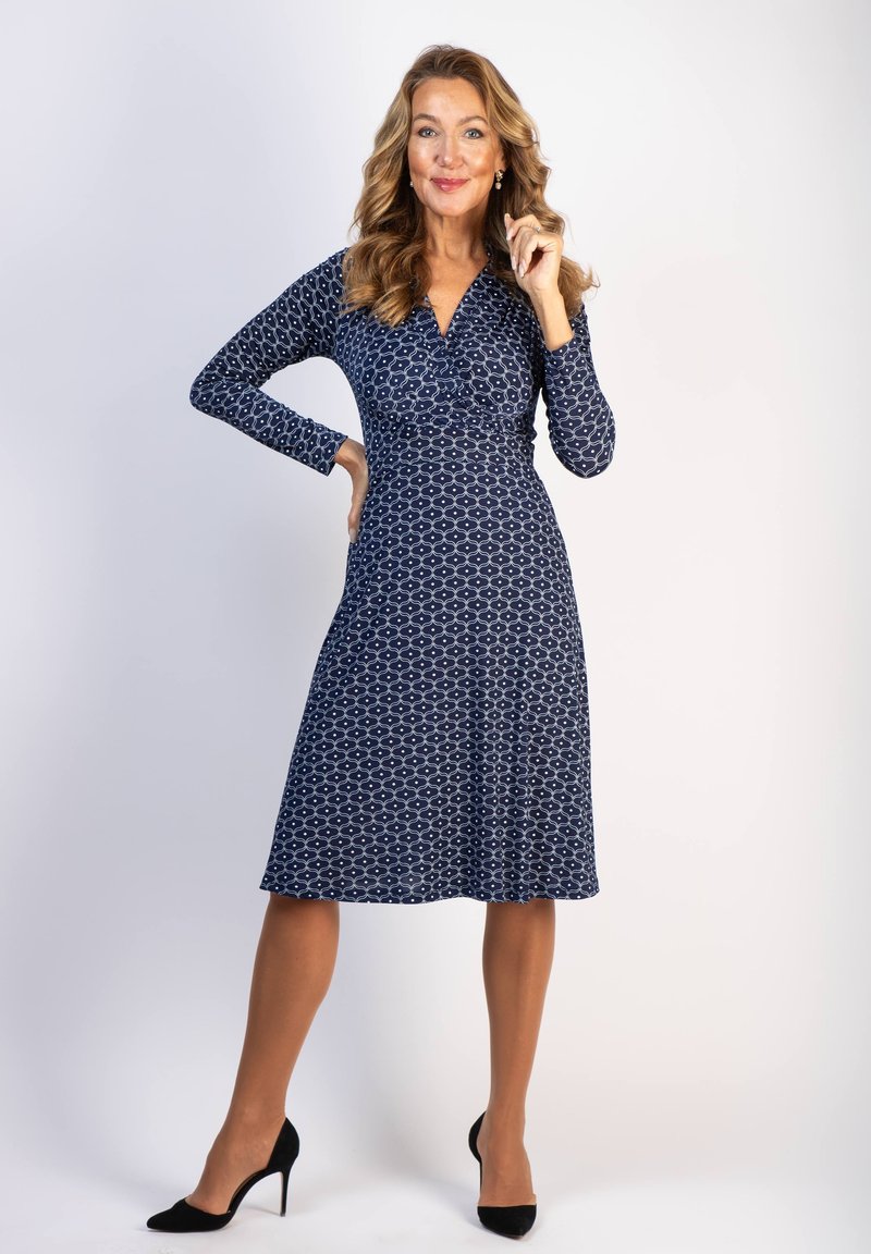 Mekko tummansinisestä kankaasta, jossa on pitkät hihat, muodikas yläosa ja leveähelma. Siinä on valkoinen geometrinen kuvio ja V-pääntie. Yhdistetty mustiin korkokenkiin.