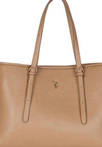 Torebka typu tote w kolorze brązowym z dwiema długimi rączkami, złotymi akcentami i małym wytłoczonym logo z przodu. Gładka faktura, minimalistyczny design.