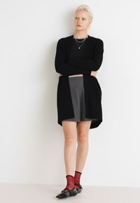 Cardigan nero indossato sopra una maglia nera, gonna plissè grigia, calzini rossi a fantasia e scarpe nere. Design minimalista con colori neutri.