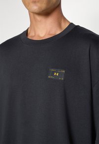 Crna pamučna majica s okruglim izrezom, s pravokutnom žutom i crnom etiketom na lijevoj strani prsiju koja sadrži logo i tekstualne detalje.