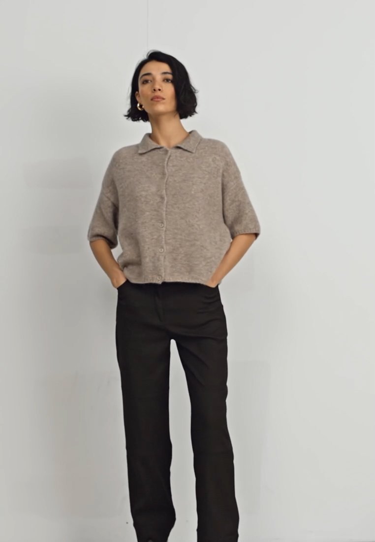 Cardigan court, en marron clair, à manches courtes, avec fermeture à boutons et col, associé à un pantalon noir taille haute, le tout sur un fond uni.