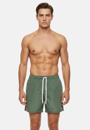 Uomo muscoloso in piedi, rivolto in avanti, indossa pantaloncini da bagno verdi con piccoli motivi di uccelli colorati e un laccio bianco.