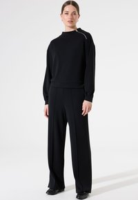 Sudadera negra con cuello alto y detalle de cremallera; combinada con pantalones negros sueltos con diseño de pliegues; confeccionada con tejido suave.