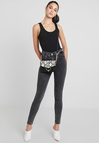 Sac bandoulière noir avec rabat en imprimé serpent, quincaillerie dorée et bandoulière enchaînée. Le mannequin porte un haut noir ajusté et un jean gris foncé avec des talons blancs.