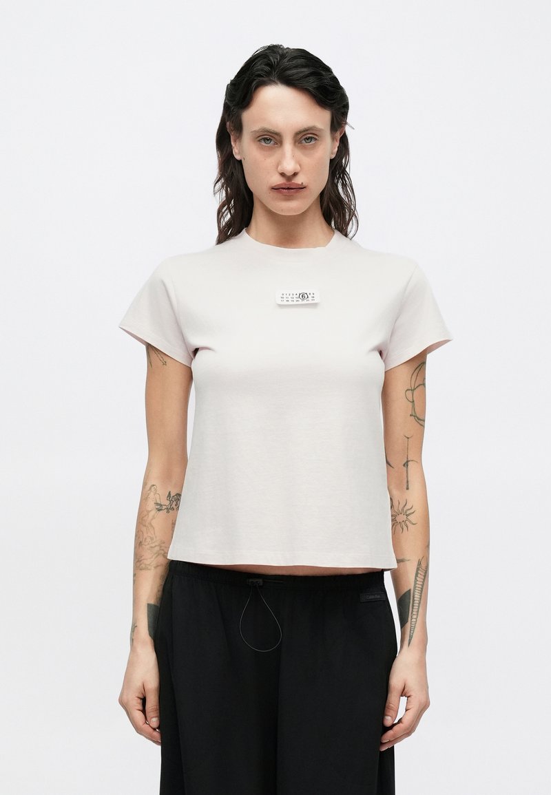 Vaaleanpunainen lyhythihainen T-paita, jossa pyöreä pääntie ja suorakaiteen muotoinen etiketti rinnassa. Kangas näyttää sileältä ja pehmeältä.