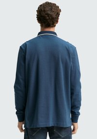 Langärmliges Poloshirt in Dunkelblau, mit strukturierter Oberfläche, offenem Kragen und kontrastierenden Abschlüssen am Halsausschnitt. Jeans sind darunter sichtbar.