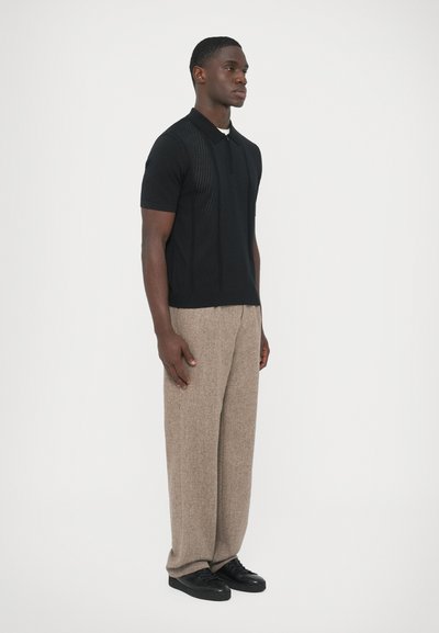 Polo preto de manga curta com padrão texturizado, combinado com calças castanho claro, largas. Modelo em pé contra um fundo branco.