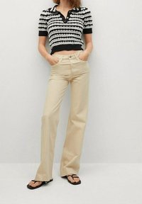 Femme portant un haut en maille rayé noir et blanc à manches courtes, un pantalon beige taille haute  à jambes larges, et des sandales noires en tongs, les mains dans les poches.