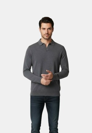 Uomo che indossa una polo grigia a maniche lunghe e jeans scuri, in piedi davanti a uno sfondo chiaro e uniforme.