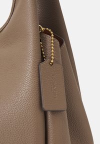 Gros plan d’un sac à main en cuir taupe texturé avec une étiquette assortie attachée par une chaîne à billes dorée.