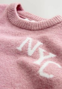 Rosa Wollpullover mit geripptem Ausschnitt und weißer "NYC" Schrift. Weiche Textur mit leicht flauschigem Finish. Nahaufnahme.