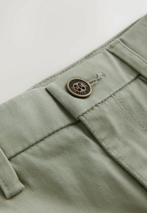 Jasnozielone materiałowe spodnie z brązowym guzikiem z odciskiem łapy i napisem "AUTENTYCZNY OBOK." Widoczne szwy i detale tekstury.