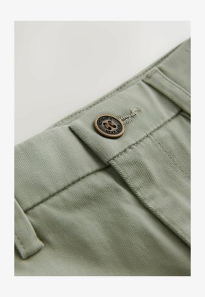 Pantaloni din țesătură verde deschis, cu un nasture bronzat care prezintă o amprentă de lăbuță și textul "AUTENTIC NEXT DOOR". Detalii vizibile de cusut și textură.