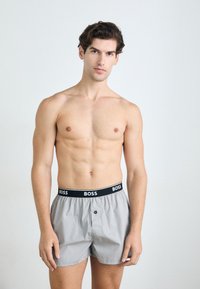 Boxers gris avec une ceinture noire portant le logo "BOSS". Le matériau semble lisse, avec un détail de bouton à l'avant.