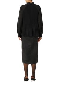 Schwarze, plissierte Bluse mit langen Ärmeln, rundem Ausschnitt und geknöpften Manschetten, kombiniert mit einem figurbetonten schwarzen Rock und Absatzschuhen mit Riemen hinten.