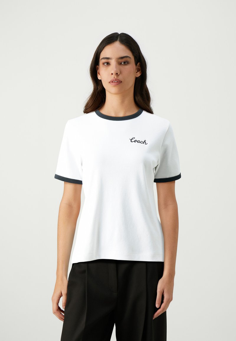White cotton t-shirt with navy ribbed collar and sleeves. Features cursive "Coach" logo on the left chest. Simple, classic design.