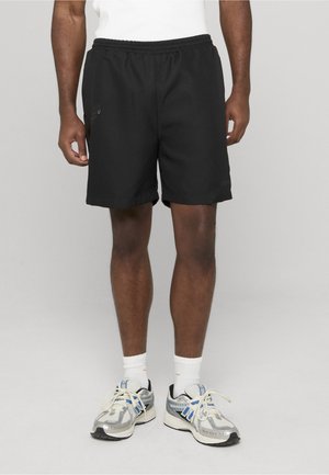 Mies, joka pukeutunut mustiin urheilushortseihin, valkoisiin sukkiin ja hopeisiin sekä sinisiin lenkkareihin, seisoo yksinkertaisen harmaan taustan edessä.