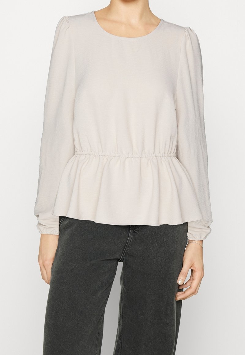 Femme portant un chemisier beige clair à manches longues avec une taille froncée et un pantalon noir taille haute, debout devant un fond blanc.