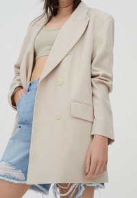 Beige dvouřadový blazer s velkými knoflíky, přiléhavý střih a dvě boční kapsy, v kombinaci s vysokým pasem a roztrhanými džíny šortkami.