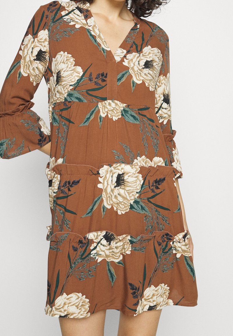 Femme portant une robe marron à fleurs blanches et à feuilles vertes, avec des manches trois-quarts et un col en V.