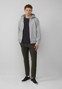 Grijze zip-up hoodie van een katoenmix, met kangoeroe-plaatsen, een trekkoordkap, gecombineerd met een donker t-shirt en olijfgroene broek, afgemaakt met zwarte sneakers.