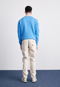 Lyseblå sweatshirt med rund halsudskæring, kombineret med cremede bukser. Bemærkelsesværdigt design inkluderer en lille lap på bukserne og struktureret stof.
