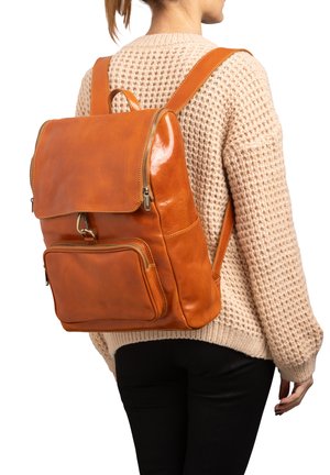 Tan leren rugzak met een glanzende afwerking, ritssluiting, voorzak en verstelbare banden, gedragen over een beige gebreide trui.