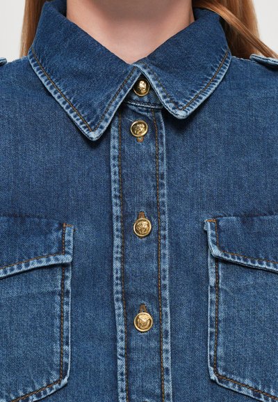 Denimskjorte med en mørkeblå nyanse, med klassisk krage, gullknapper og to brystlommer med klaffelukking.