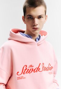 Rózsaszín kapucnis pulóver az elején piros hímzéssel "The Stwd Studio" felirattal, kék-fehér csíkos inggel rétegezve.