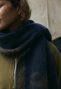 Ciemnoniebieski szalik wykonany z miękkiego, teksturowanego materiału, owinięty wokół szyi, z nutą jaśniejszego koloru wtkanego w tkaninę.
