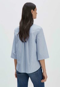 Camicia a righe blu con maniche a tre quarti, tessuto morbido, orlo curvo e sottili righe multicolori, indossata con jeans blu.