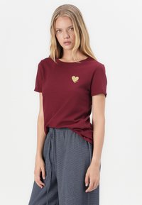 Mladá žena s dlhými blond vlasmi, oblečená v vínovom tričku so zlatým srdcovým emblémom a sivých voľných nohaviciach, stojaca na bielom pozadí.