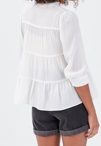 Blouse blanche texturée avec volants à étages et manches trois-quarts, associée à un short en denim foncé avec revers retroussés.