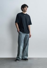 Jeune homme aux cheveux bouclés portant un t-shirt noir oversize avec le texte "OFF", un jean large bleu clair avec des coutures et des baskets noires, debout contre un mur blanc.