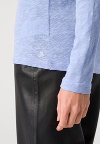 Svijetloplava majica dugih rukava od teksturiranog materijala, u kombinaciji s crnim kožnim hlačama. Mali logo prikazan na rubu.