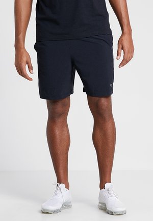 Mann in schwarzen Sportshorts, schwarzem T-Shirt und weißen Turnschuhen, der vor einem schlichten hellen Hintergrund steht.