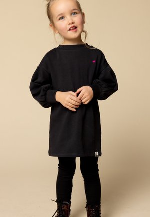 Zwarte tuniek met lange mouwen en plooien bij de mouwen, heeft een klein roze hartlogo op de borst. Gecombineerd met zwarte leggings en gebloemde laarzen.