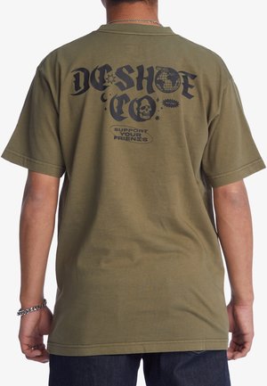 Man wearing olive green T-shirt with black graphic text "DC SHOE CO" and skull, globe, and stars printed on the back.