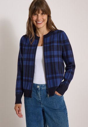 Femme souriante portant un cardigan zippé à carreaux bleu et noir, une chemise blanche et un jean bleu avec une main dans la poche, sur un fond uni.