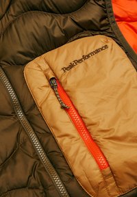 Detail einer grünen und orangen Daunenjacke mit gestepptem Design, einer hellbraunen Reißverschlusstasche mit dem Logo "Peak Performance" und einem kontrastierenden orangenen Reißverschluss.