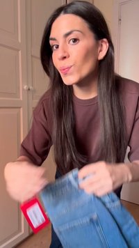 Γυναίκα με μακριά σκούρα μαλλιά που φοράει καφέ πουκάμισο κρατά ένα ζευγάρι μπλε τζιν με κόκκινη ετικέτα τιμής σε ένα δωμάτιο με κλειστές λευκές πόρτες.